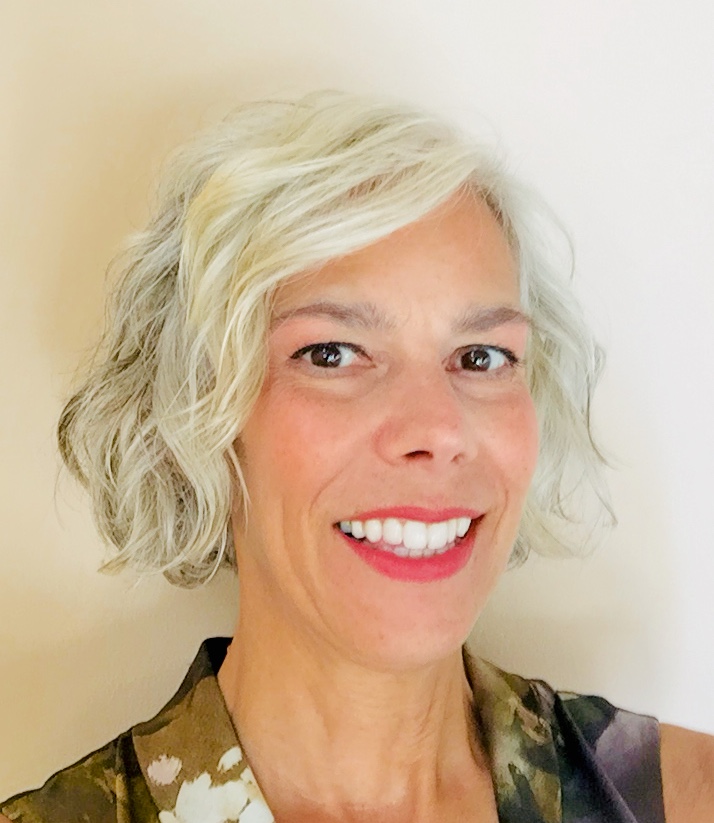 Headshot of Lori standing against a white wall. A white woman with short gray hair wearing bright pink lipstick.