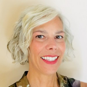 Headshot of Lori standing against a white wall. A white woman with short gray hair wearing bright pink lipstick.