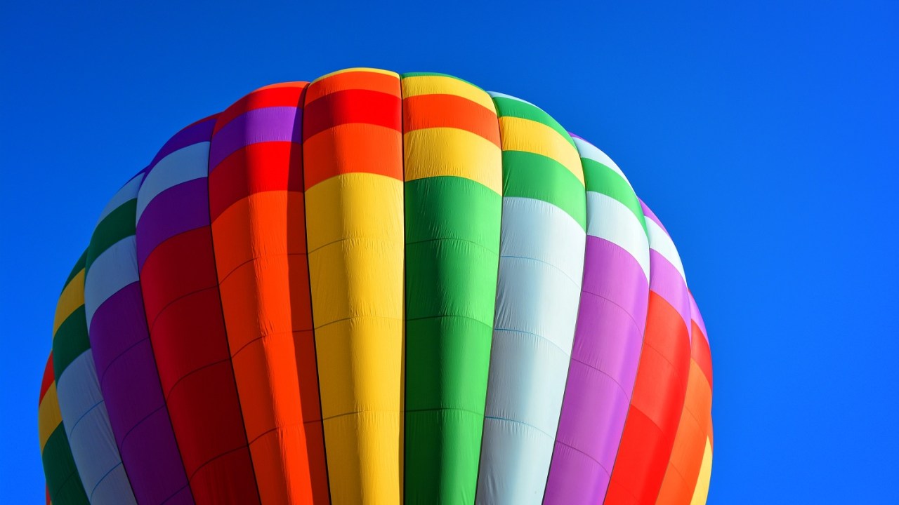 Closeup of a rainbow colored hot air balloon floating against a clear blue sky.