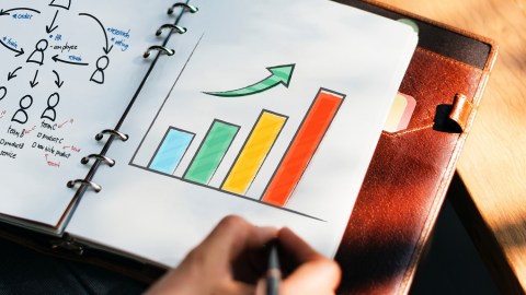 An open leather journal with someone drawing a bar graph in different colors.