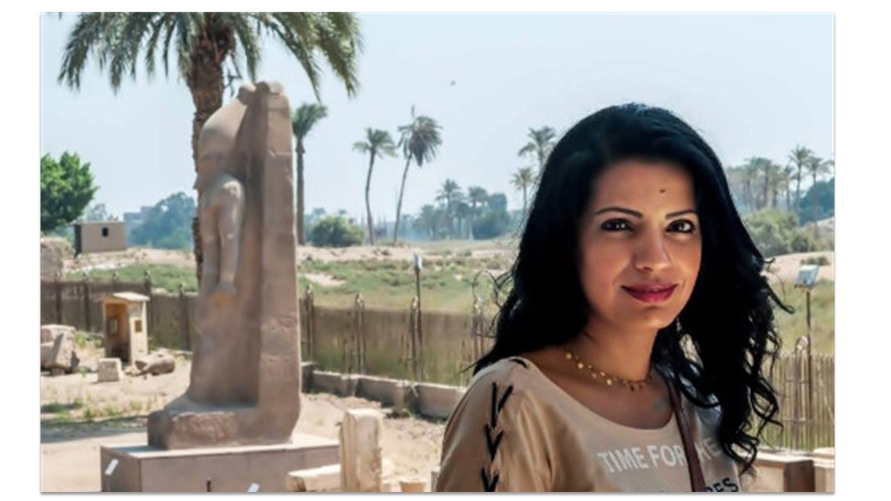 An Egyptian woman stands to the right in front of some Egyptian ruins.