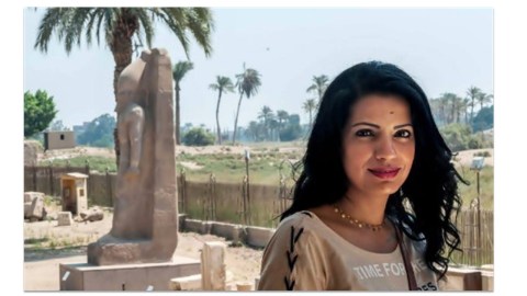 An Egyptian woman stands to the right in front of some Egyptian ruins.