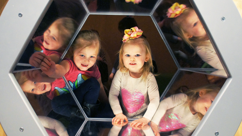Two children peek through a mirrored hexagonal window.