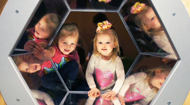 Two children peek through a mirrored hexagonal window.