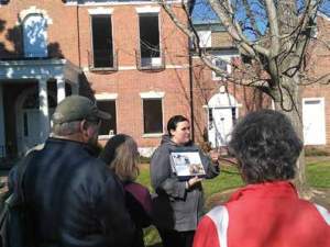 A young white woman stands in front of a group of people outside the front of a red brick brick building holding a placard which appears to be a map.
