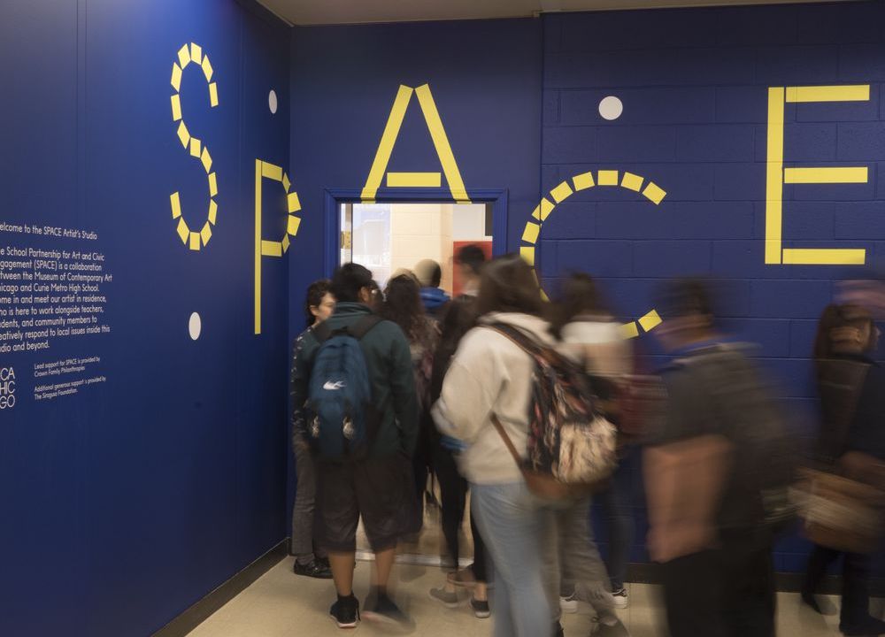 A group of students wearing coats and backpacks stand in line to enter through a door in a dark blue wall with the letters S P A C E written in yellow.