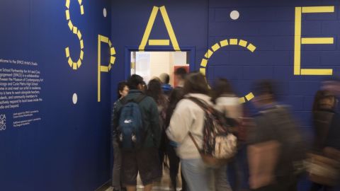 A group of students wearing coats and backpacks stand in line to enter through a door in a dark blue wall with the letters S P A C E written in yellow.
