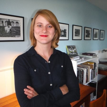 A white woman stands in a room with framed pictures on the wall behind her. She stands with her arms crossed.