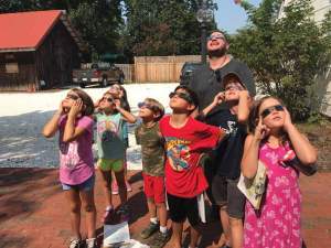 A group of young children an done adult look up at the sun wearing solar glasses.