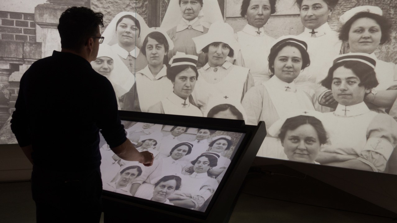 A visitor to the International Center of Photography is seen viewing a historical black-and-white photo of a group of women in uniform. The photo appears on the screen in front of the visitor, which he touches, and in larger size in front of him. Using the touch screen, he is able to change his view of the photograph.