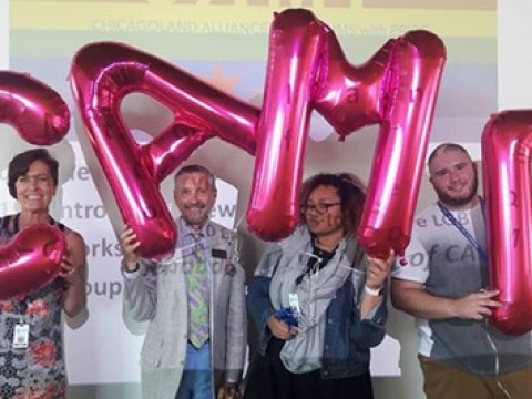 Four members of CAMP are seen holding large, hot pink mylar balloons above their heads, one letter for each person, which spell out CAMP.