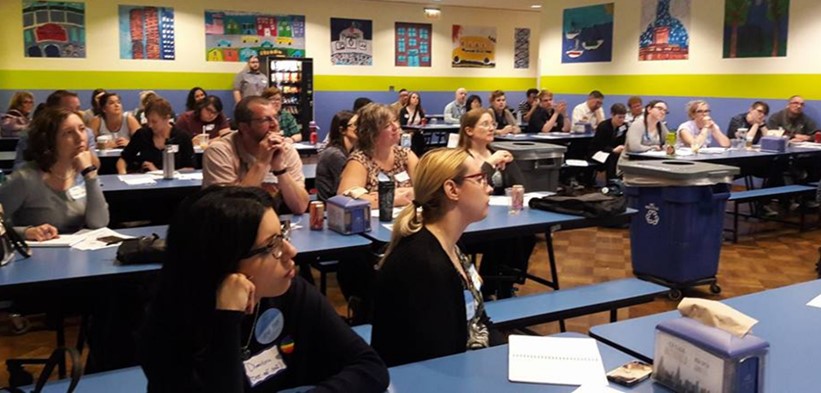 A large number of attendees at a CAMP meeting are shown with their attention facing toward the front of a room, where a presentation on LGBTQ-friendliness in museums is being given