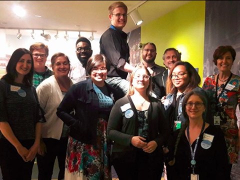 12 members of the CAMP group working to make museums more LGBTQ-friendly pose for a group photo indoors. Some wear badges and name tags. One appears to stand on a ladder or stepstool.