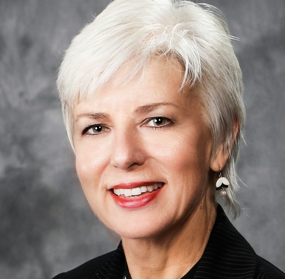 Headshot of a white woman smiling at the camera with short cut white hair wearing a black striped suit jacket and white and black shirt.