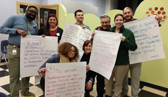 A group of eight individuals participating in CAMP hold up signs with sentences written on them proposed to be included in the group's mission statement, designed to make museums more friendly and inclusive to LGBTQ people.