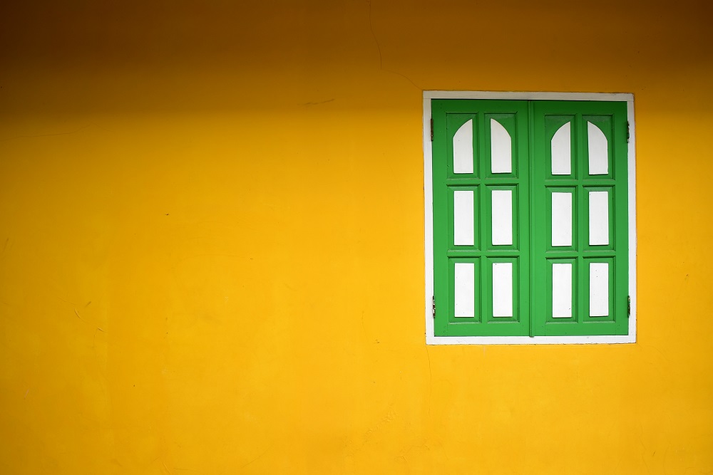 View of a yellow painted wall with a shuttered window painted in green and white off-center on the wall.