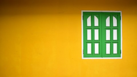 View of a yellow painted wall with a shuttered window painted in green and white off-center on the wall.