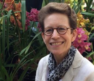 View of a woman standing in front of a garden scape smiling wearing horn rimmed glasses and a white jacket and blue and white scarf.