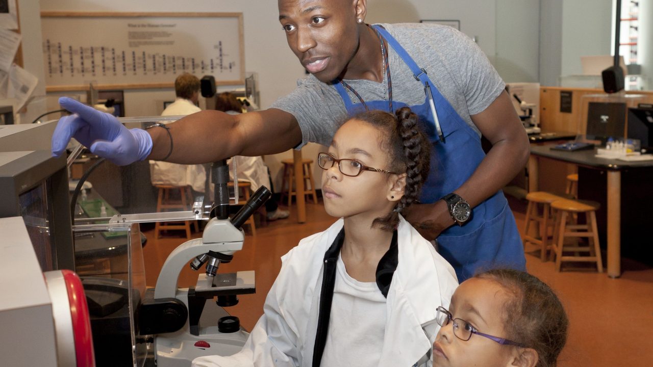In the Kitty Andersen Youth Science Center, an instructor wearing an apron and gloves points to a display, while two child pupils study it with him, wearing lab coats and safety gloves. A microscope and other lab equipment sits on the desk in front of them.