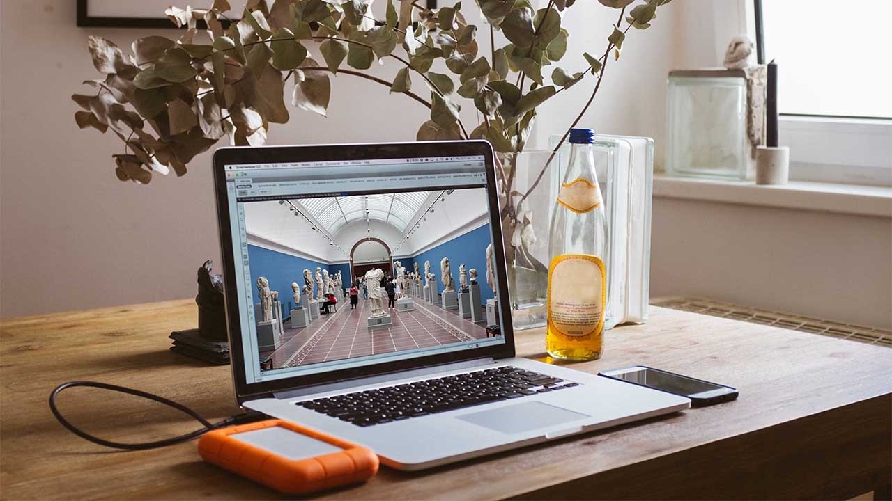 Closeup of a desk with an open laptop and flowers in a vase with a bottle of orange liquid.