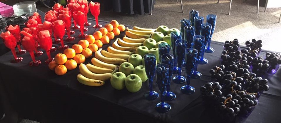The rainbow-color-coordinated fruit spread at CAMP's kickoff meeting is pictured, with red watermelon, orange oranges, yellow bananas, green apples, blue blueberries, and purple grapes arranged into consecutive stripes of color corresponding to the rainbow.