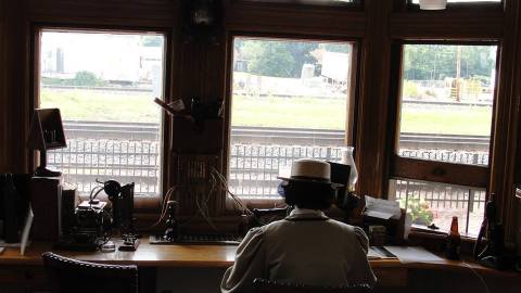 A woman sits at a desk wearing period costume. On the desk is an exhibition of several historical telegraphs. Railroad tracks are visible in the window in front of her.
