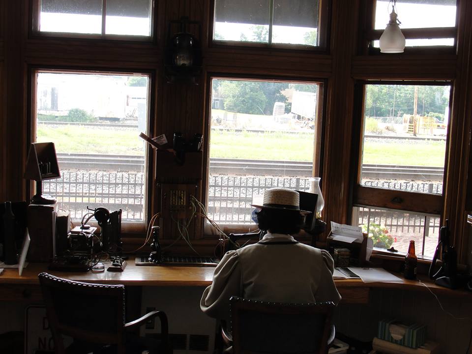 A woman sits at a desk wearing period costume. On the desk is an exhibition of several historical telegraphs. Railroad tracks are visible in the window in front of her.