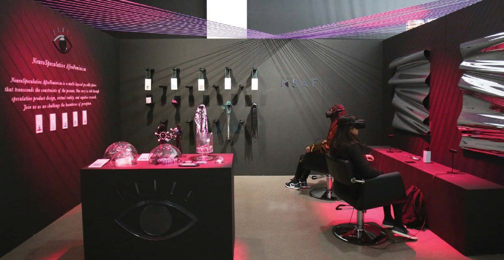 Two people sit in chairs facing different directions wearing immersive googles in a dimly lit space with a table and things hanging on the walls around them.