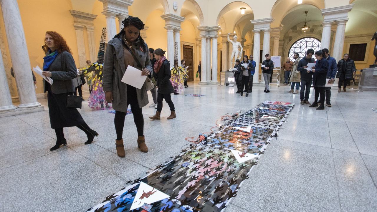 A young black woman stands in a museum inspecting a flat printed piece that is displayed on the floor.