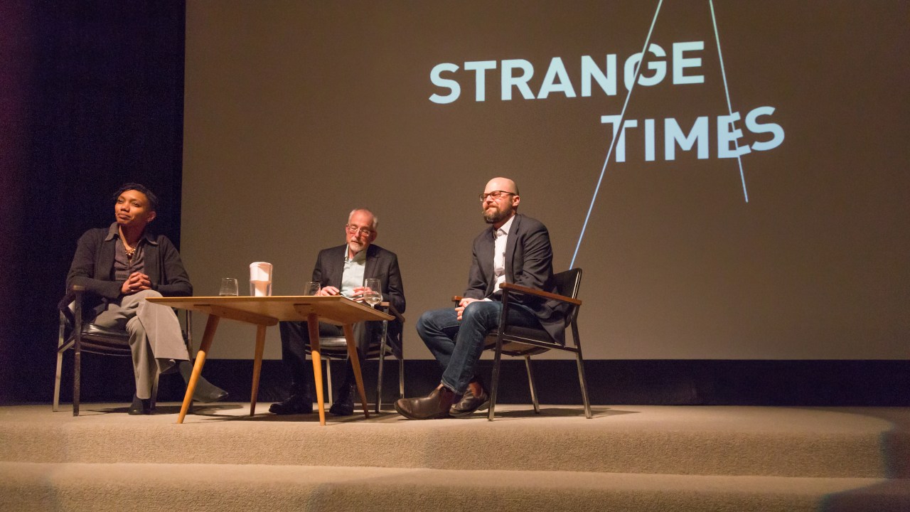 Speakers sit around a table in front of a screen with the name of the program series, "Strange Times"