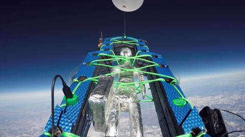 In a rendering, a sculpture of a human figure covered with shiny silver metallic material like tin foil is strapped into secure restraints and attached to a big pink balloon. It floats high into the sky over the ground below.
