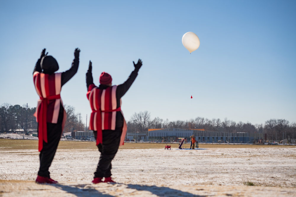 Two people wearing matching outfits face away from the camera and toward the weather balloon in the distance, which is beginning its ascent to space.
