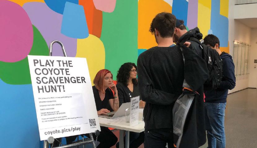 Several young men stand at a table with their backs to the camera where two women sitting down are talking to them with an easal erected to the left hand side that says "Play the Coyote Scavenger Hunt".