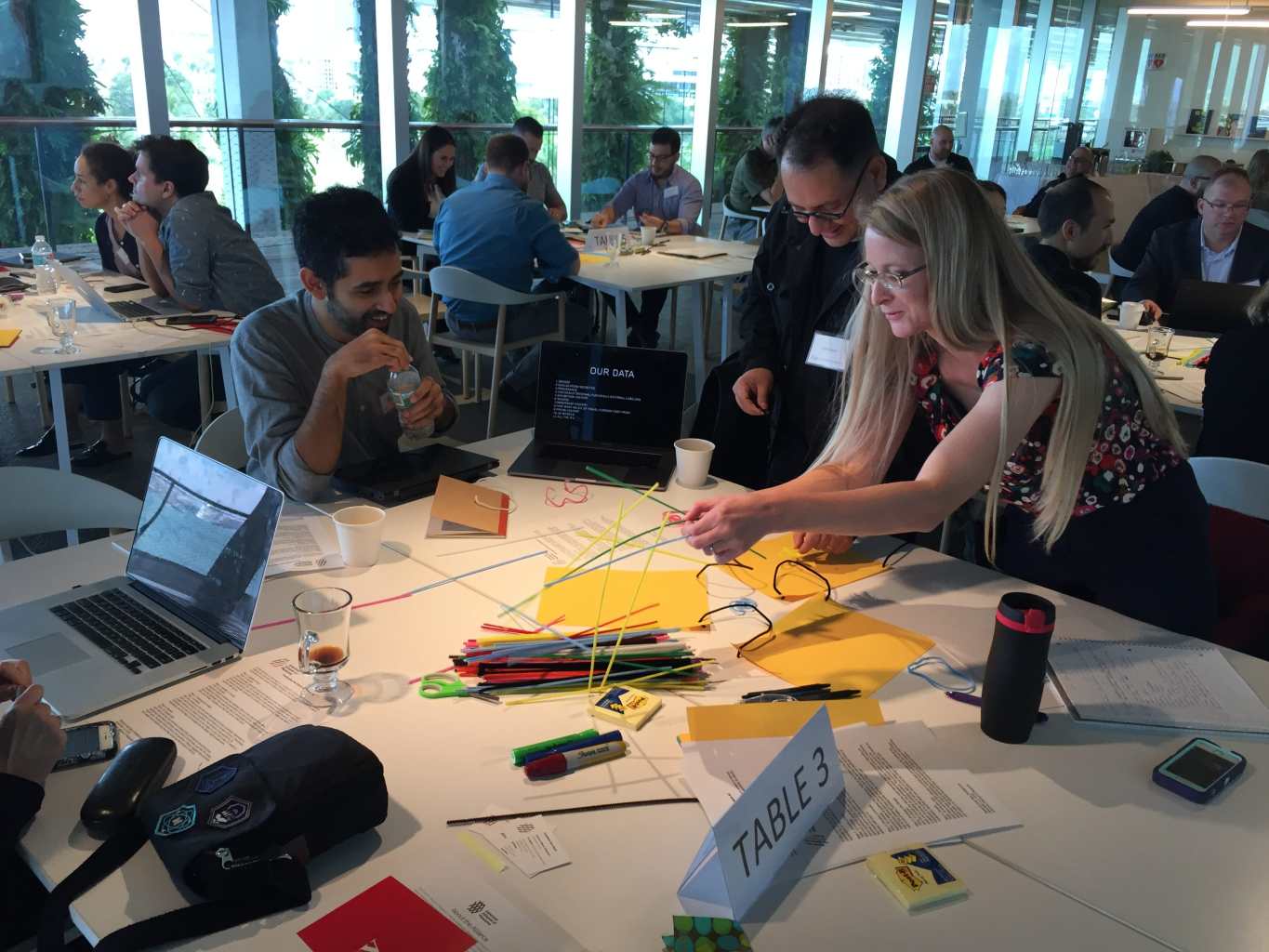 A group of people gathers around a table looking at the pipe cleaners and pieces of paper they had to make their prototype with. Two of the people are in the process of building the prototype by connecting the pieces of paper with the pipe cleaners. A laptop screen is visible showing a PowerPoint presentation with a slide titled "Our Data."