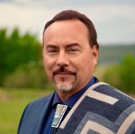 Headshot of a dark-haired man standing outside with a goatee wearing a geometric style blanket on one shoulder and a bolo tie.