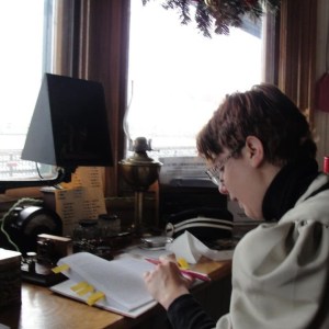 A white woman with short brown hair sits at a desk with a pencil in her let hand looking at book on the desktop which is situated under a set of windows.