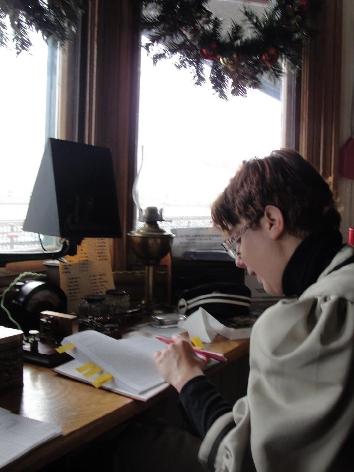 A white woman with short brown hair sits at a desk with a pencil in her let hand looking at book on the desktop which is situated under a set of windows.