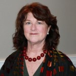 Image of a white woman with shoulder length wavy reddish brown hair wearing a large red beaded necklace.