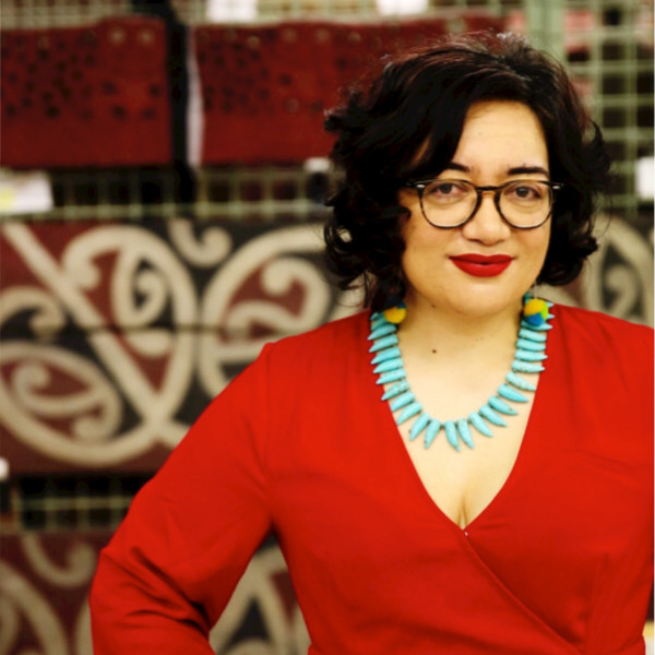 Professional shot of Puawai Cairns a dark haired woman wearing dark rimmed glasses, a red surplice top and turquoises colored necklace standing in front of a decorative tile wall.