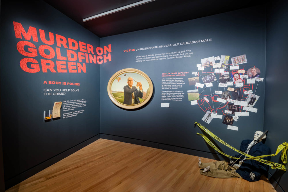 In a gallery space, an experimental exhibition game called "Murder on Goldfinch Green" is displayed. On the floor is a skeleton model dressed in a men's suit and posed with a golf club, and cordoned off with caution tape. The walls have a photograph of a man playing golf and a diagram showing the connections between evidence and suspect photographs with string. The visible text on the wall reads: "A body is found! Can you help solve the crime? Use this card as a guide to find these artworks and read the cards next to them for evidence. Check off those artworks that you don't think were involved or write notes along the side. Come back to The Niche for the reveal to prove your detective skills. Victim: Charles Chase, 65-year-old Caucasian male. Chase was a well-to-do banker who loved to golf. The manner of death has been ruled a homicide, but we are waiting for more definite results from Professor Rand. Cecelia Chase reported her husband missing after he didn't return home from golfing last Saturday. Police could find no lead, but a psychic suddenly appeared at the Chase home, telling them to look for sand and the sound of finches. This tips them to Goldfinch Green Golf Course where the body was found in a shallow grave. This is a now deserted gold course, since the new Country Club has opened."
