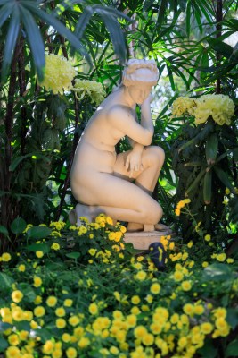 A white marble statue of a nude female figure kneeling is displayed inside of a garden, with green foliage and yellow flowers surrounding it.