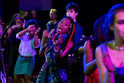 The performer dances and sings on stage wearing a sunburst-print dress and cowrie jewelry. Other performers join in on the dancing behind her.