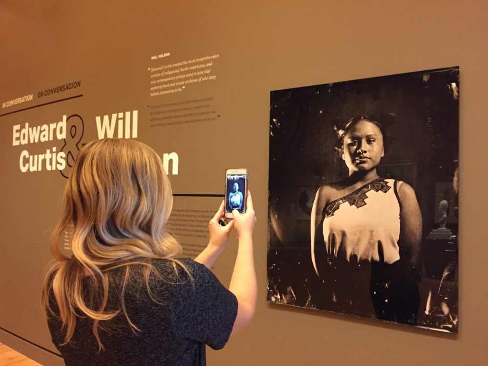 In an exhibition gallery a visitor stands in front of a photograph of a Native American woman. The photograph is rendered on the visitor's phone screen.
