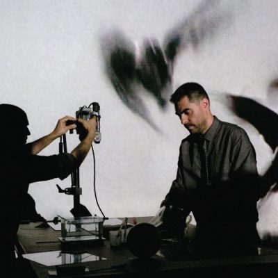 Two men stand on stage in front of a desk with equipment on it. Behind them is a projection of birds in flight.