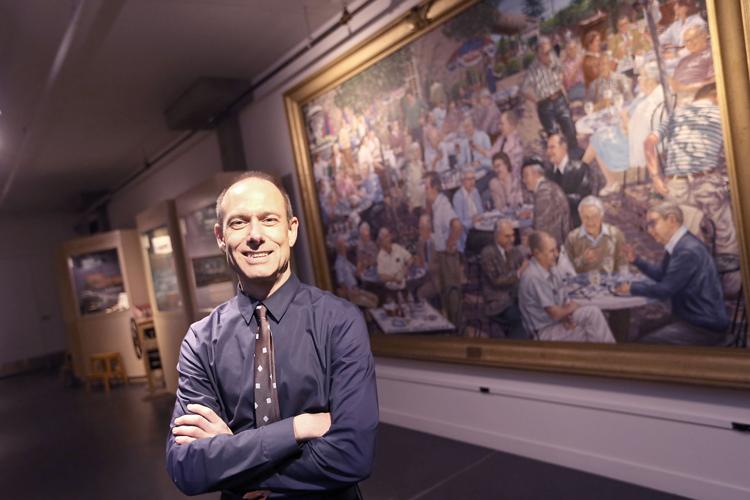 Headshot of a white man with short grey hair standing wiht his arms crossed standing in front of an oil painting framed in an ornate gilt frame.