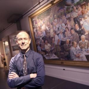 Headshot of a white man with short grey hair standing wiht his arms crossed standing in front of an oil painting framed in an ornate gilt frame.