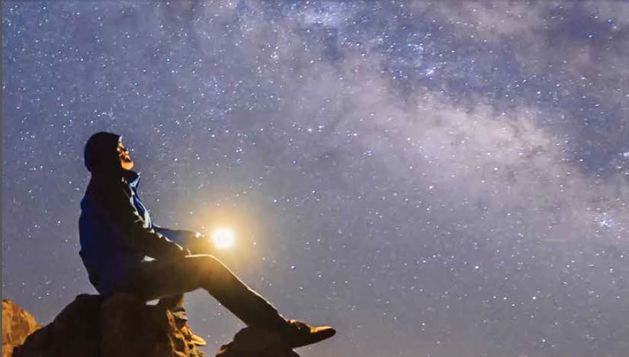A man sits on a large rock at dusk staring up at the stars shining brightly above.