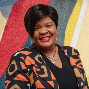 Image of a black woman with straight short black hair standing in front of a colorful backdrop, smiling and wearing a geometric patterned jacket.