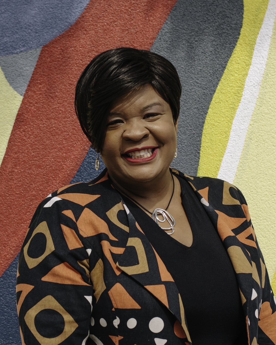 Image of a black woman with straight short black hair standing in front of a colorful backdrop, smiling and wearing a geometric patterned jacket.