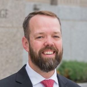 Headshot of a white man with a full beard and mustache smiling at the camera standing outside near a large wall wearing a suit and red tie.